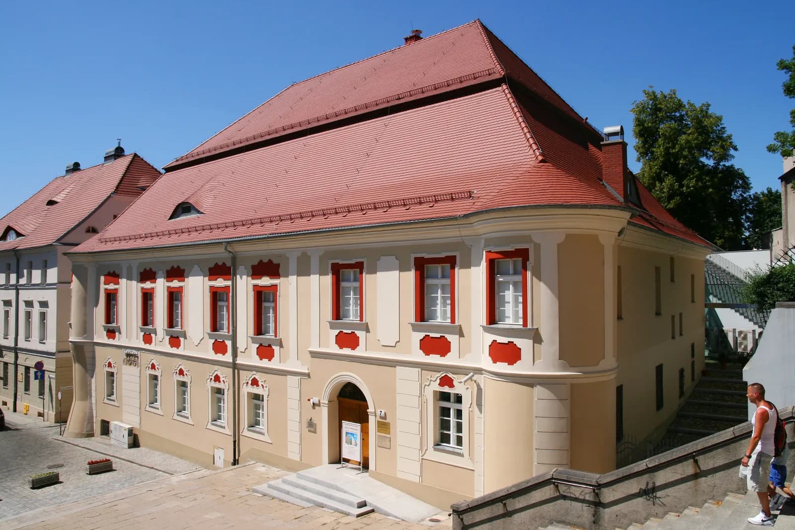 A large building with a red tile roof and light-colored walls, featuring red-framed windows, a stepped entrance, and a slight curve to its facade, with a person walking down steps on the right.
